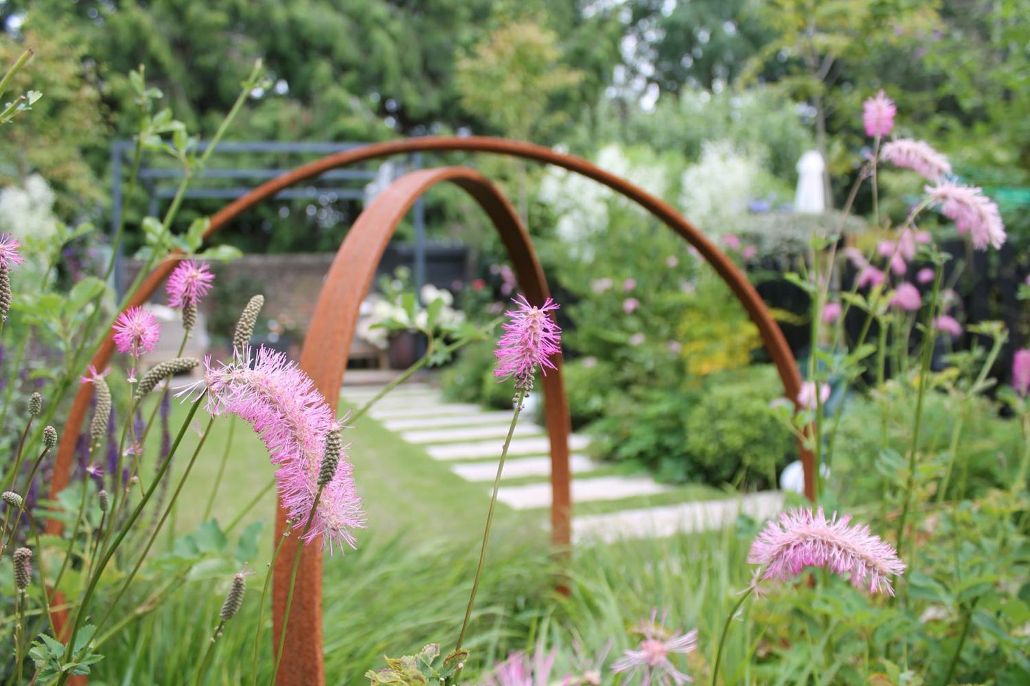Weathered steel arches standing in a loose, prairie-inflected border of Sanguisorba and grasses — structure that gives the planting something to grow against and the garden something to read in winter.