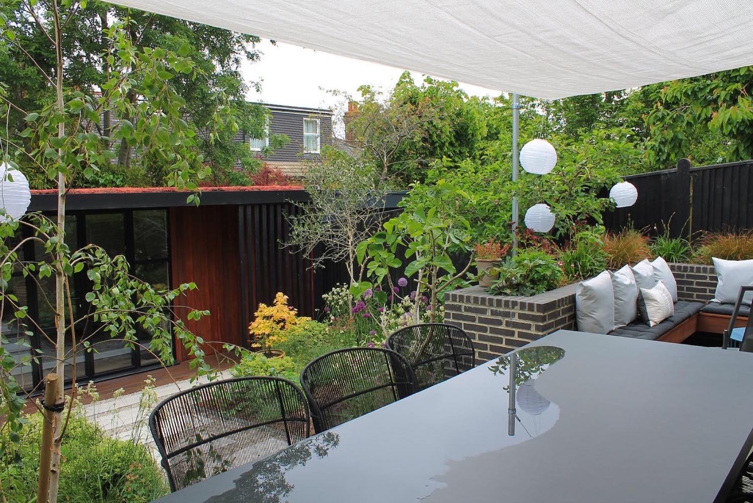 A contemporary dining terrace under a tensioned canopy, with built-in brick and timber seating, dark-stained boundaries and lanterns hung through the planting so the space still works after dark.